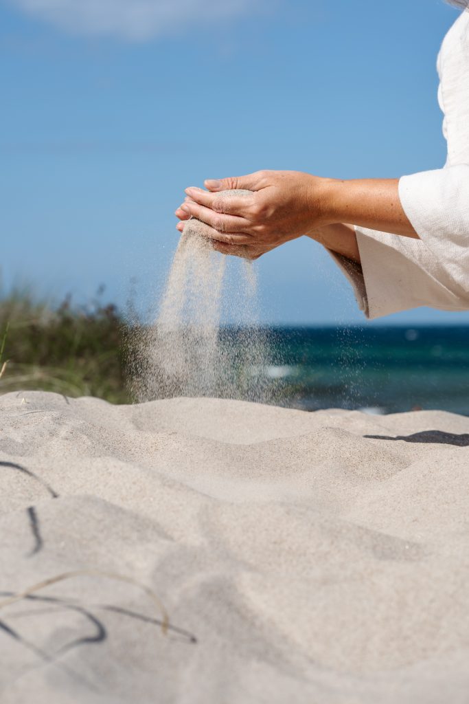 Sand rinnt durch Marlens Hände, im Hintergrund Dünen und Meer